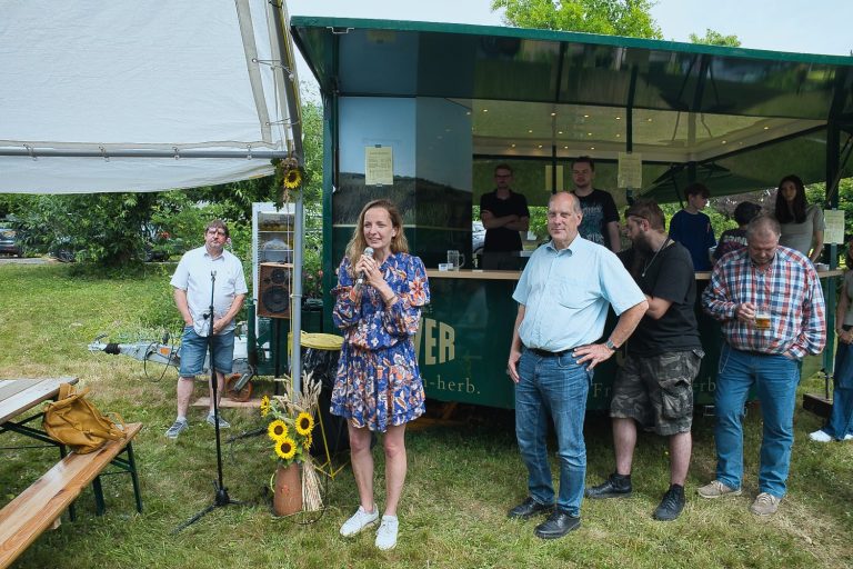 Menschen stehen vor einem Verkaufsstand auf einem Fest, in einer grünen Umgebung.