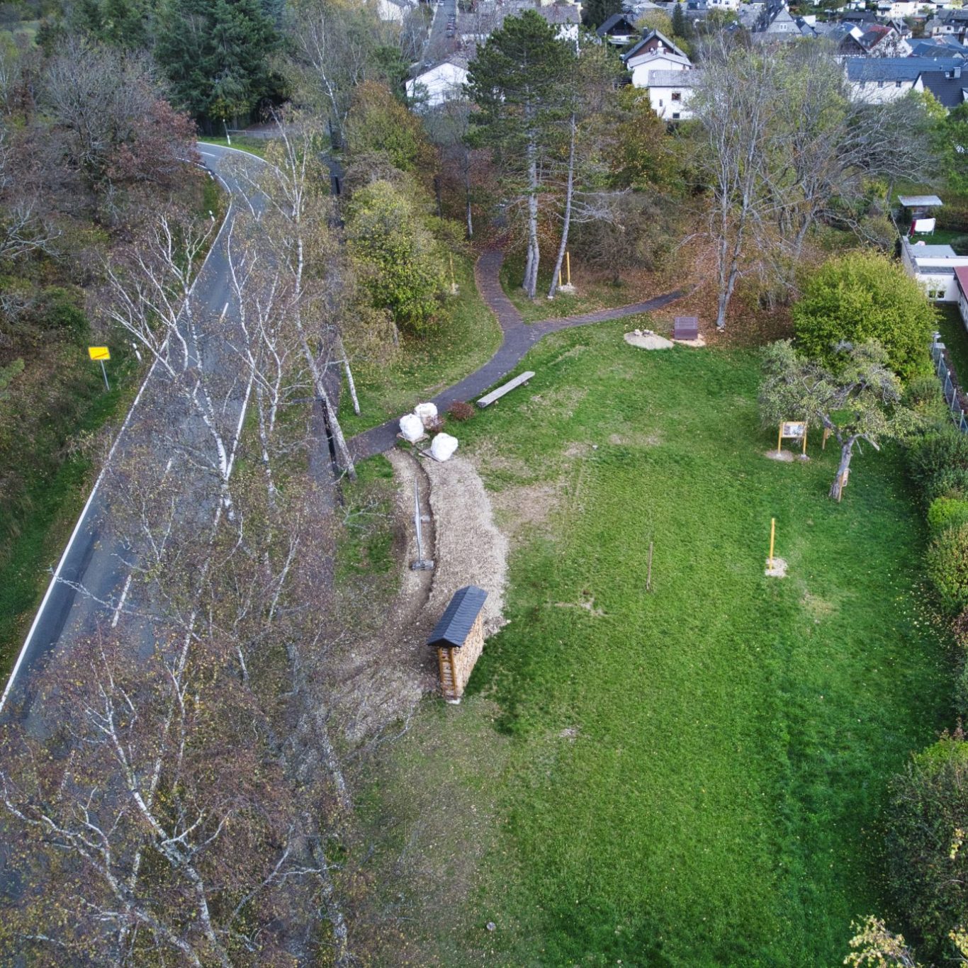 Luftaufnahme eines Parks mit Wegen, Bäumen und Spielgeräten neben einer Straße.